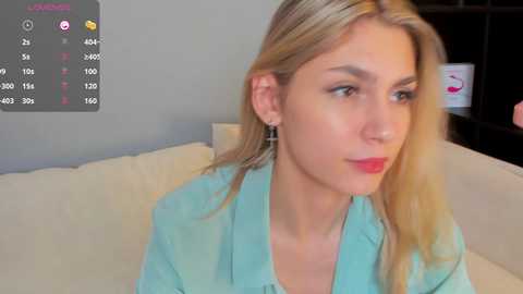 Media: Video of a blonde Caucasian woman with fair skin, wearing a light blue shirt, looking contemplative, sitting on a beige sofa. Background includes a grey wall and a black shelf with a white mug.