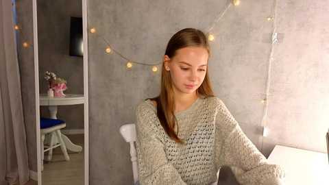 Media: Video of a young Caucasian woman with straight brown hair, wearing a loose-knit gray sweater, sitting indoors with a gray wall, string lights, and a small table in the background.