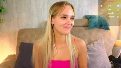 Media: A video of a smiling, fair-skinned, blonde woman with straight hair, wearing a pink tank top, sitting on a beige sofa with grey and teal pillows, in a cozy, well-lit living room.