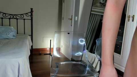 Media: Video of a minimalist bedroom with a clear acrylic chair in front of a white wardrobe, reflecting light, and a neatly made bed with white sheets in the background.