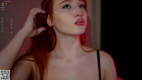 Media: Video of a young woman with fair skin and long, straight red hair, wearing a black top, standing in a dimly lit room with a red light.