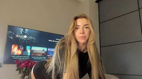 Media: Video of a blonde woman with long hair, wearing a black shirt, in a modern living room with a TV showing \"Game of Thrones\" and a vase of red flowers.