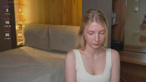 Media: A video of a blonde woman with fair skin, wearing a white tank top, sitting in a modern bedroom with wooden furniture, a TV showing a virtual reality scene.