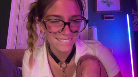 Media: Video of a smiling woman with glasses, wearing a white shirt and black choker, sitting indoors with a braided ponytail, dim lighting, and a framed picture in the background.