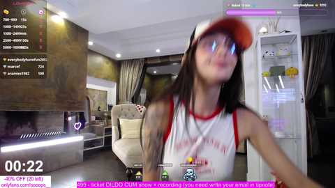 Media: Video of a slender, tattooed woman with long dark hair wearing a red hat and white tank top, holding a bottle in a modern, dimly lit living room.