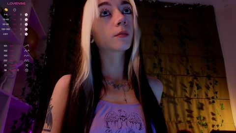 Media: Video of a young woman with pale skin, long black and blonde hair, tattoos, and piercings, wearing a white tank top, standing in front of a dark, vine-covered wall, with a dimly lit, moody room background.