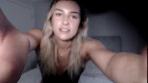 Media: Video of a young woman with long, blonde hair and fair skin, wearing a black top, reaching out with her arms towards the camera, taken in a dimly lit, simple bedroom.