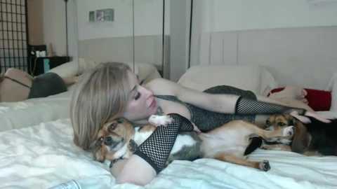 Media: Video of a blonde woman in black fishnet stockings, lying on a white bed with a small dog and a beagle. Background includes white walls and a wardrobe.