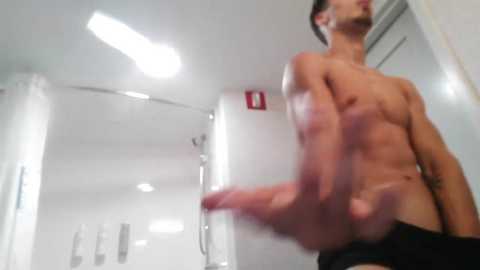 Media: Video of a shirtless, fit man with short brown hair, wearing black briefs, pointing his erect penis towards the camera. Background shows a modern, white-tiled bathroom with a glass shower door and a red emergency exit sign.