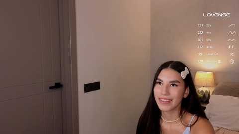 Media: Video of a smiling, light-skinned Latina woman with long, dark hair, wearing a white hair clip and a light blue top, standing indoors next to a calendar and a lit lamp.