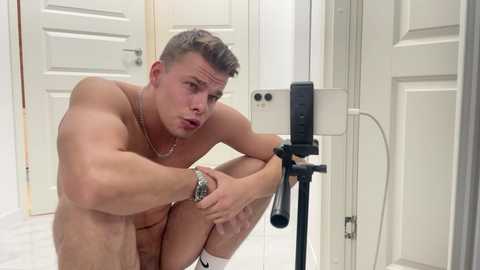 Media: Video of a fit, shirtless man with short, light brown hair, holding a microphone, sitting on a white floor in a minimalist room with white doors and a camera stand.