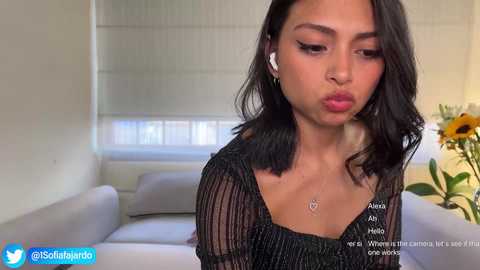 Media: Video of a young woman with medium skin tone, black hair, and a black mesh top, pouting in a modern living room with white sofa, sunflower, and white blinds.