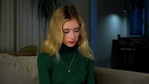 Media: A video of a young Caucasian woman with blonde, wavy hair, wearing a green ribbed turtleneck and a silver necklace, sitting on a beige couch in a dimly lit living room.