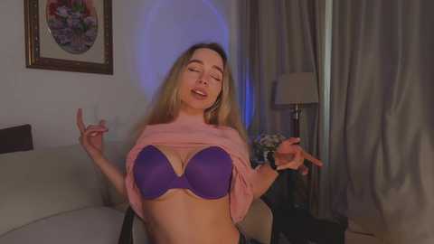 Media: Video of a blonde woman in a purple bra, lifted shirt, making peace signs, in a dimly lit room with framed art and a lamp.