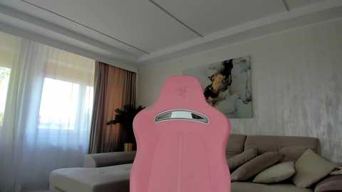 Media: Video of a modern living room with a pink gaming chair, beige sofa, abstract wall art, sheer curtains, and natural light streaming in.