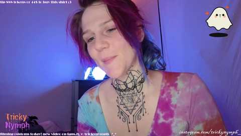 Media: Video of a young woman with dyed purple hair, wearing a tie-dye shirt, and a large, intricate black tattoo of a dreamcatcher on her neck. She is indoors with a blue-tinted wall and a glowing lamp in the background.