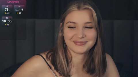 Media: Video of a smiling, fair-skinned woman with straight, shoulder-length blonde hair, wearing a black strapless top, against a dark background.