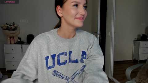 Media: Video of a smiling young woman with fair skin and brown hair in a ponytail, wearing a gray UCLA sweatshirt. She is seated indoors, with a white dresser and vase of flowers in the background.