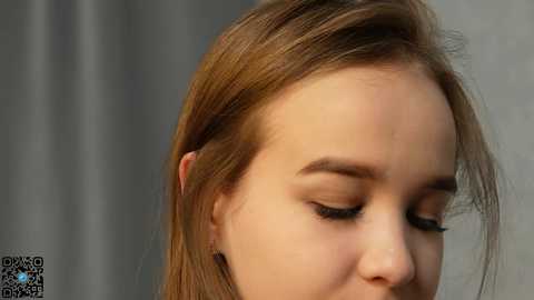 Media: Close-up video of a young woman with light skin and straight, shoulder-length brown hair, eyes closed, resting on her right cheek. Background is blurred, featuring vertical, light-colored, possibly metal or wooden, vertical panels.