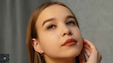 Media: Video of a young Caucasian woman with light skin, straight blonde hair, and green eyes. She wears minimal makeup and has a contemplative expression. The background is a blurred, neutral tone.