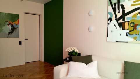 Media: A modern, minimalist living room with green and white walls, abstract art, a white couch, and a vase of white flowers on a side table.