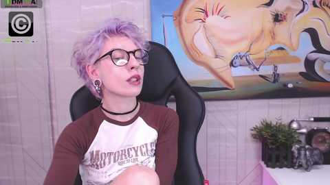 Media: Video of a pale-skinned, androgynous person with short, lavender hair, wearing black-framed glasses, a brown and white t-shirt, and a black choker, sitting in a black gaming chair. The background features a vibrant, abstract painting and a small plant.