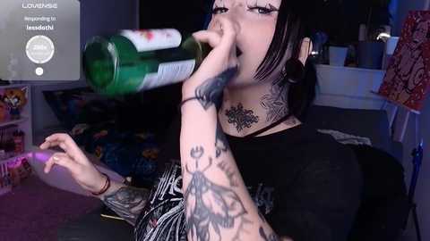 Media: Video of a tattooed woman with black hair, drinking beer, wearing a black shirt, in a dimly-lit room with a cluttered background.