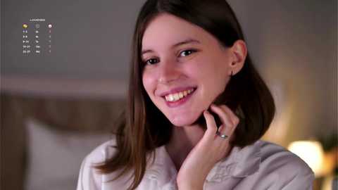 Media: Video of a smiling woman with shoulder-length brown hair, wearing a white robe, against a blurred, softly lit indoor background.