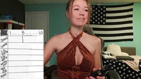 Media: Video of a young, fair-skinned, blonde woman in a brown halter top, seated in a gaming chair in a room with a large American flag and a whiteboard with numbers and words.