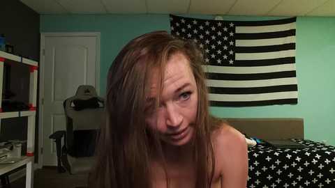 Media: A video of a young woman with long brown hair, wearing a black tank top, leaning forward in a room with a white door, black and white American flag, and a cluttered desk.