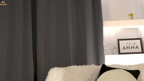 Media: Video of a modern bedroom with dark grey curtains, white wall, and a framed \"Anna\" sign. A plush white pillow and a black and white decorative pillow rest on a fluffy white blanket.