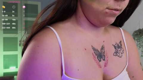 Media: A video of a plus-sized woman with fair skin, long brown hair, wearing a white spaghetti-strap tank top, showcasing two butterfly tattoos on her collarbone. Background includes a window with green blinds and a white wall.