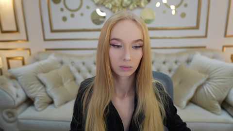 Media: Video of a blonde, fair-skinned young woman in a black shirt, standing in a plush, elegant living room with a white, tufted sofa and golden decor.