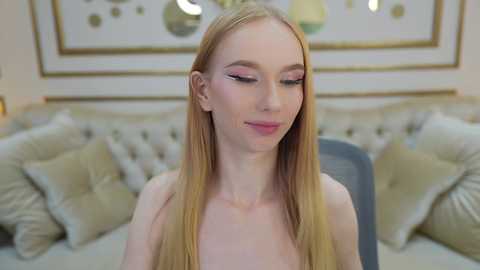 Media: A video of a fair-skinned, slender young woman with long blonde hair, wearing pink eyeshadow and a smile, sitting on a beige couch adorned with gold cushions and framed art.