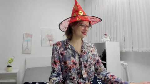 Media: Video of a young woman with fair skin, wearing a red wizard hat and floral-patterned shirt, sitting in a modern, minimalist room with white walls and a white curtain.