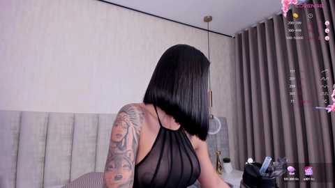 Media: A video of a woman with shoulder-length black hair and a tattoo sleeve, wearing a sheer black top, sitting on a bed with grey headboard, in a room with beige walls and grey curtains.