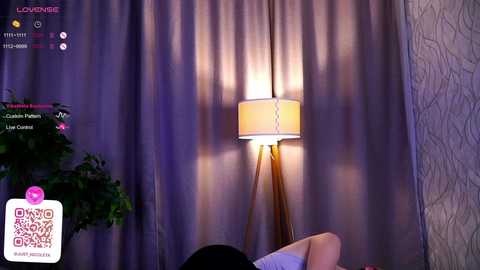 Media: A video of a woman with dark hair, wearing a white top, sitting in a room with a lamp casting a soft glow on beige curtains and a potted plant.