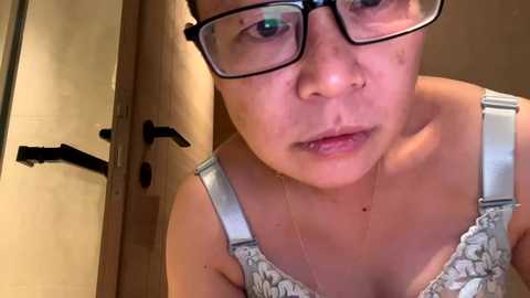 Media: Video of an Asian woman with short black hair, wearing glasses and a floral-patterned top, standing in a bathroom with beige walls and a wooden door.