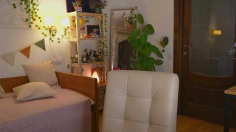 Media: Video of a cozy, well-lit bedroom with a twin bed, white chair, wooden bookshelf, and green plants; warm lighting, eclectic decor, and a wooden door in the background.