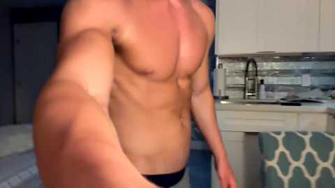 Media: Video of a shirtless, muscular man with a defined six-pack, wearing only black briefs, standing in a modern kitchen with white cabinets and a backsplash.