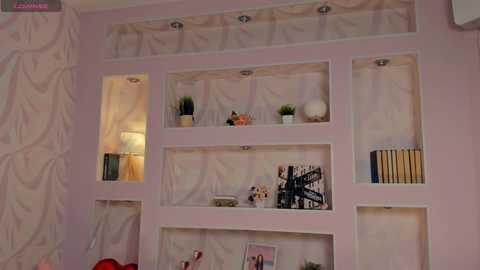 Media: Video of a pastel pink, bookshelf-lined wall with open shelving, featuring books, plants, a globe, and decorative items, creating a cozy, organized, and stylish living space.