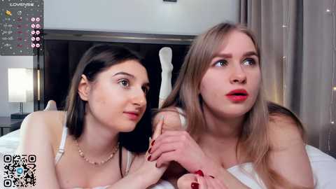 Media: Video of two young women, one with dark hair, the other blonde, lying on a bed in a modern bedroom. They hold a dildo, discussing it with serious expressions.