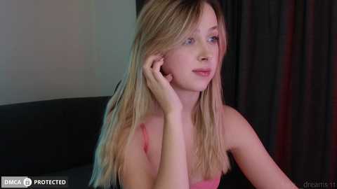 Media: A video of a young Caucasian woman with long blonde hair, wearing a pink tank top, sitting on a black sofa. She has fair skin and is looking contemplatively to the side.