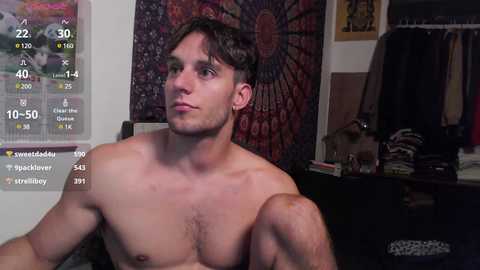 Media: Video of a shirtless, fair-skinned, fit young man with short brown hair, looking contemplative, in a dimly lit room with a dark tapestry, a calendar, and a coat rack in the background.