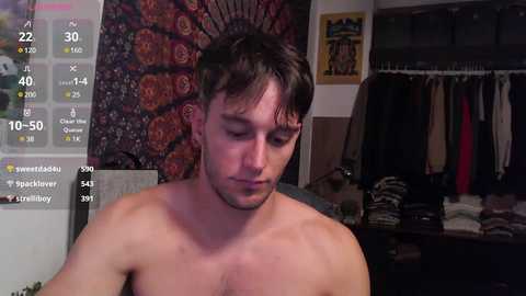 Media: Video of a shirtless, young, muscular man with short dark hair, eyes closed, standing in a dimly lit room with a colorful tapestry and hanging clothes.