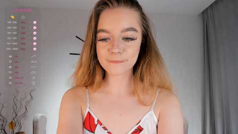 Media: Video of a young woman with light skin, long blonde hair, wearing a white and red floral camisole, smiling, in a modern bedroom.