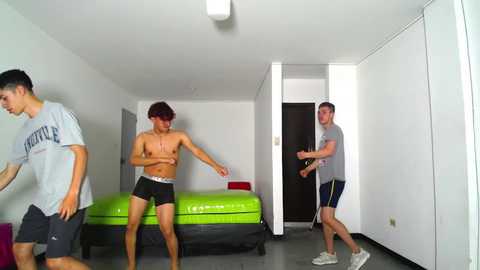 Media: Video of three shirtless young men in tight black shorts playing ping-pong in a small, white-walled room with a bright green inflatable bed and white cabinets.