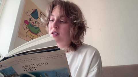 Media: Video of a young Caucasian boy with shoulder-length, wavy brown hair, reading a book titled \"\u0410\u041f\u0420\u0410\u0428\u041a\u0418\u041d, \u0415\u0422\" in a white T-shirt, sitting on a beige couch against a light beige wall with a colorful cartoon poster.