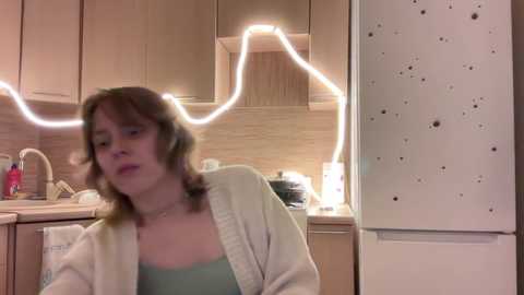 Media: Video of a young woman with shoulder-length, wavy brown hair, wearing a light green top and a white cardigan, seated in a kitchen with beige cabinets and a white refrigerator.