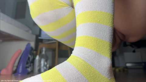 Media: A close-up video of a woman wearing yellow and white striped socks, with her breasts partially visible. Background shows kitchen items, including a bottle and purple container.